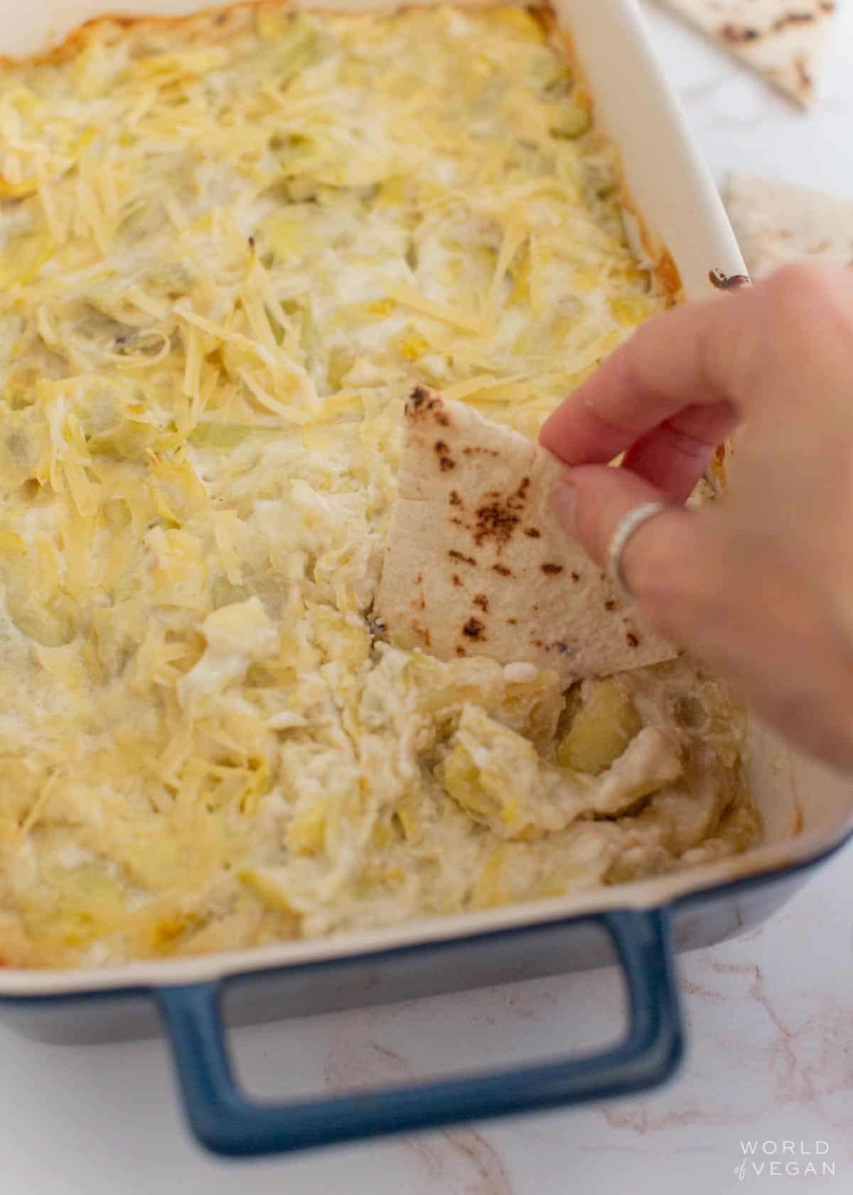 A hand dipping pita bread in a pan of vegan artichoke dip.