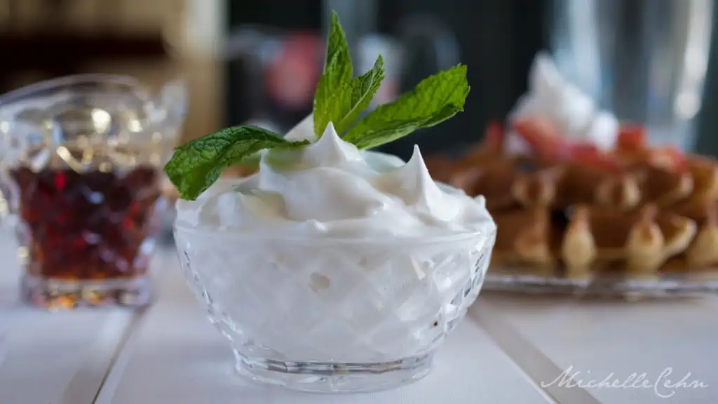 aquafaba vegan whipped cream in a glass bowl with a mint sprig garnish and pancakes and maple syrup in the background