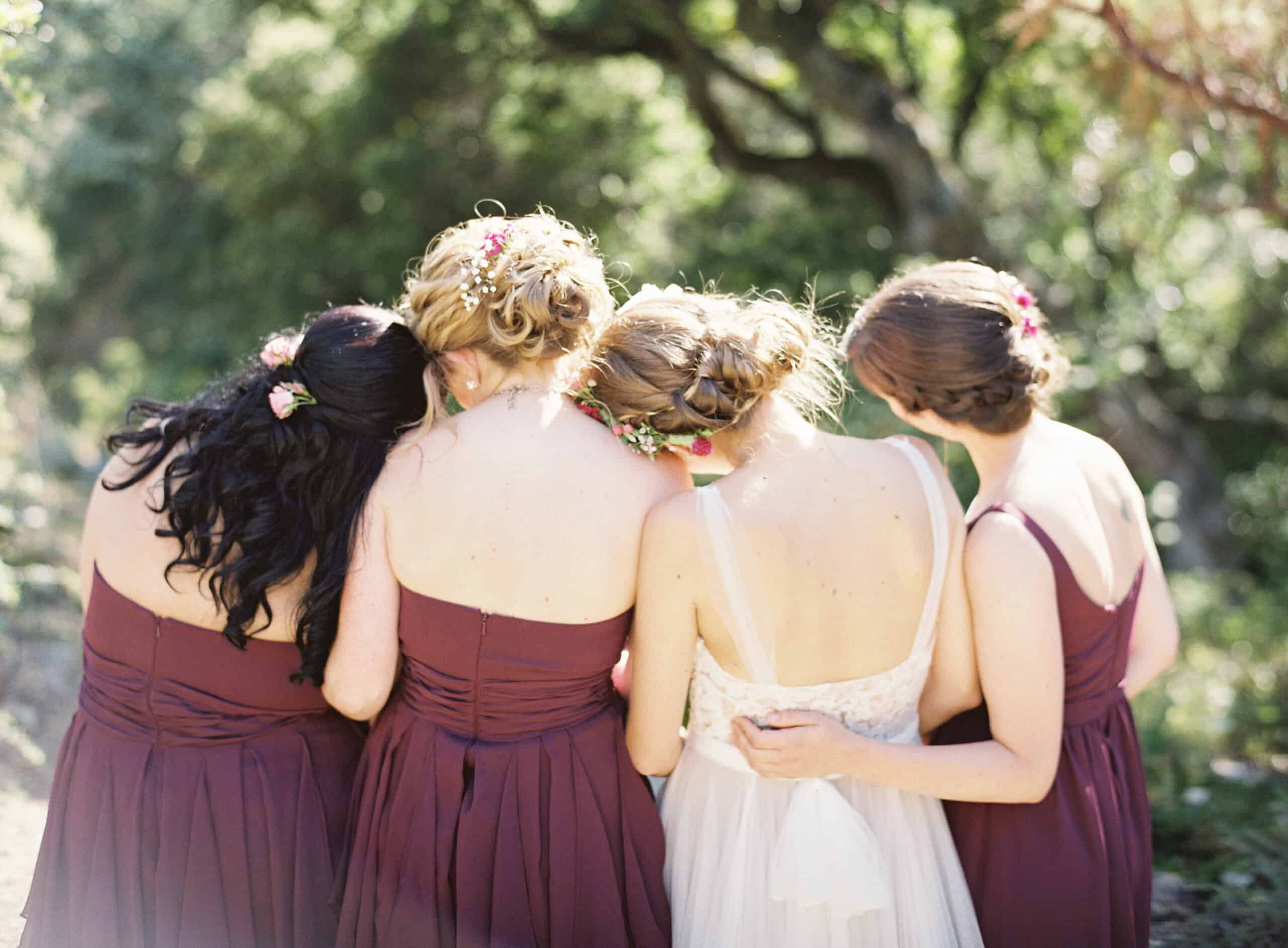 Bridal party in plum dresses hugging under dappled sunlight.