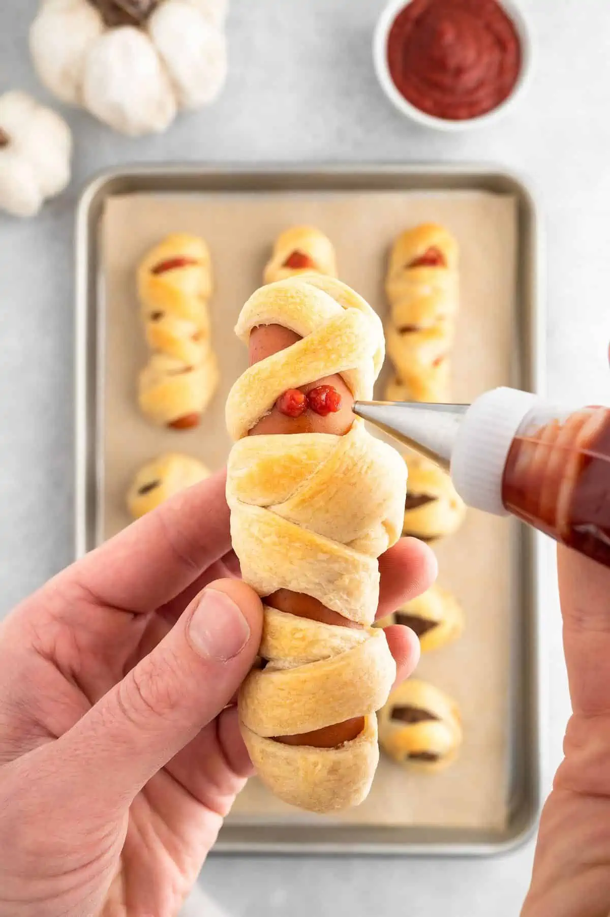 Hand holding a mummy dog while adding ketchup eyes with a piping tool.