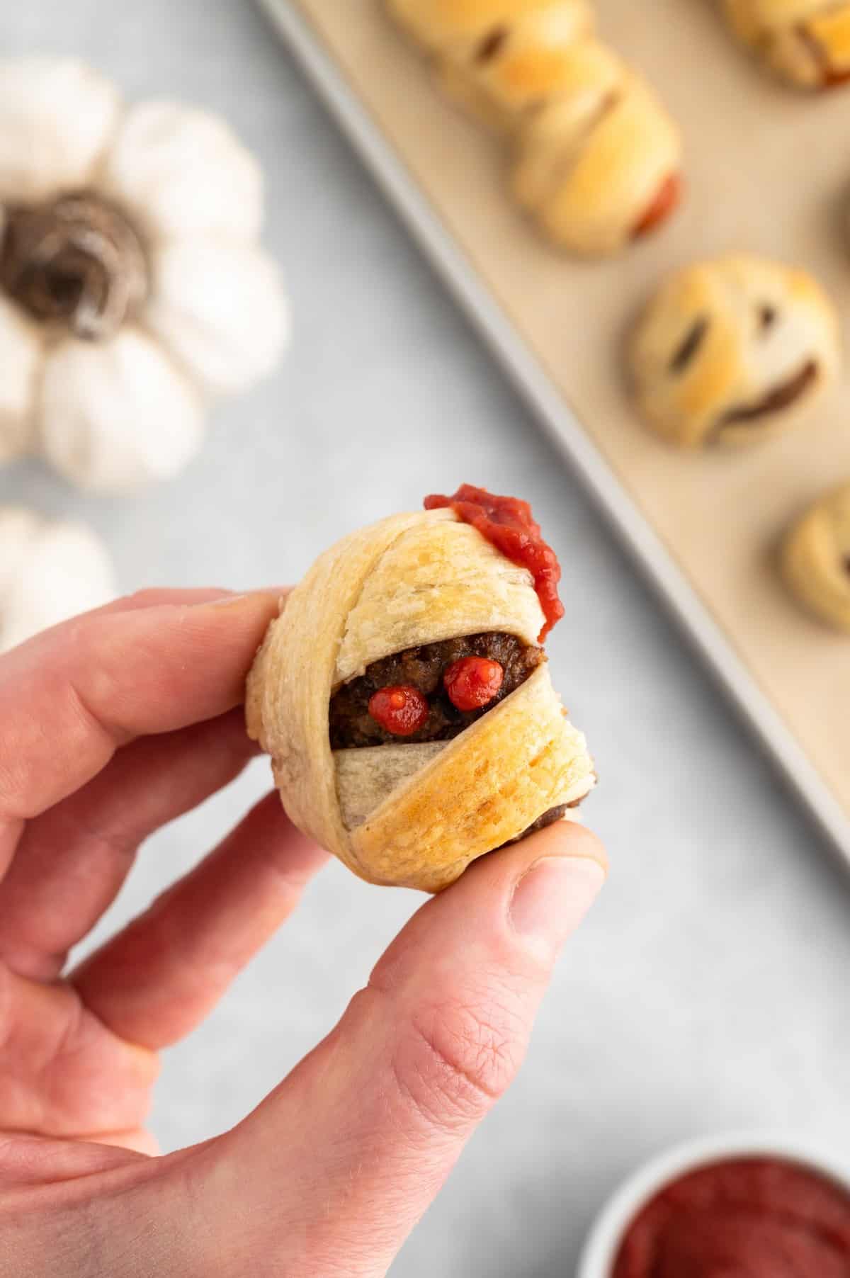 Hand holding a mummy ball that has been dipped in ketchup and a bite missing from its side.