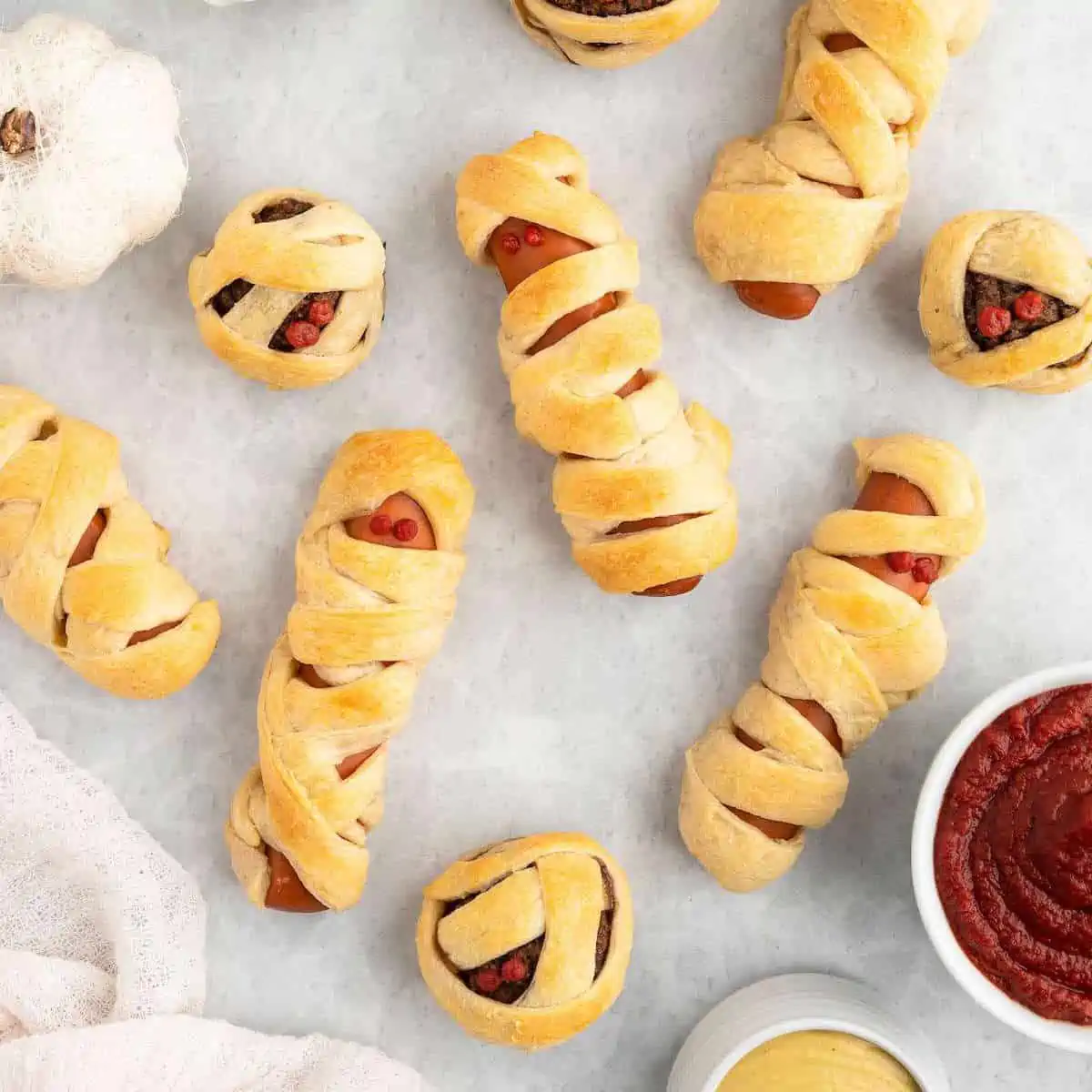Vegan mummy dogs and ball scattered on a surface with bowls of dipping sauce to the side.
