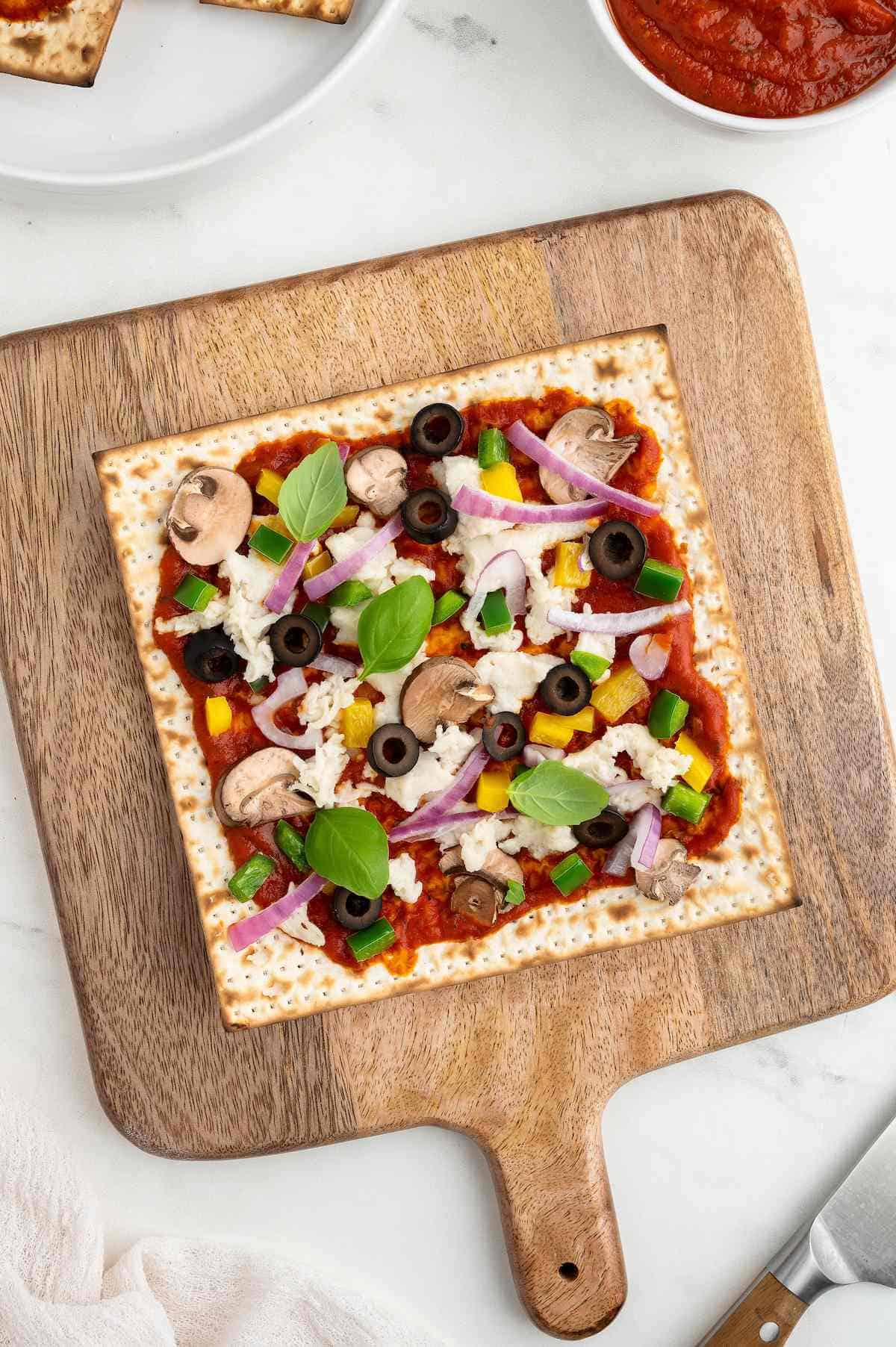 Matzah pizza topped with a variety of vegetables and vegan cheese on a wooden pizza tray.