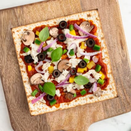 Matzah pizza topped with a variety of vegetables and vegan cheese on a wooden pizza tray.