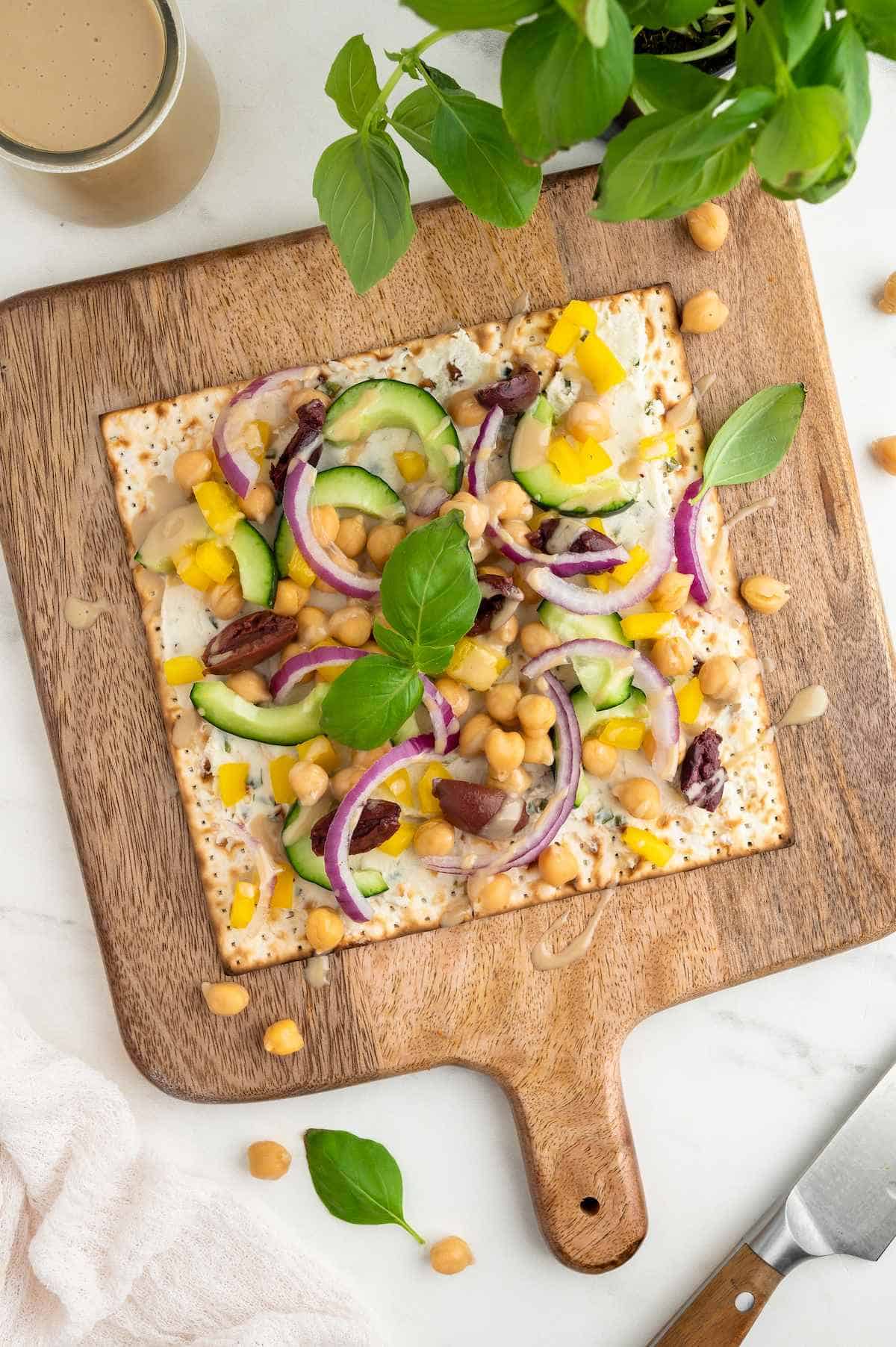 Greek style matzah pizza topped with Onion & Chive dairy-free cream cheese, chickpeas, yellow bell peppers, kalamata olives, basil, sliced cucumber, red onion, and a drizzle of tahini.