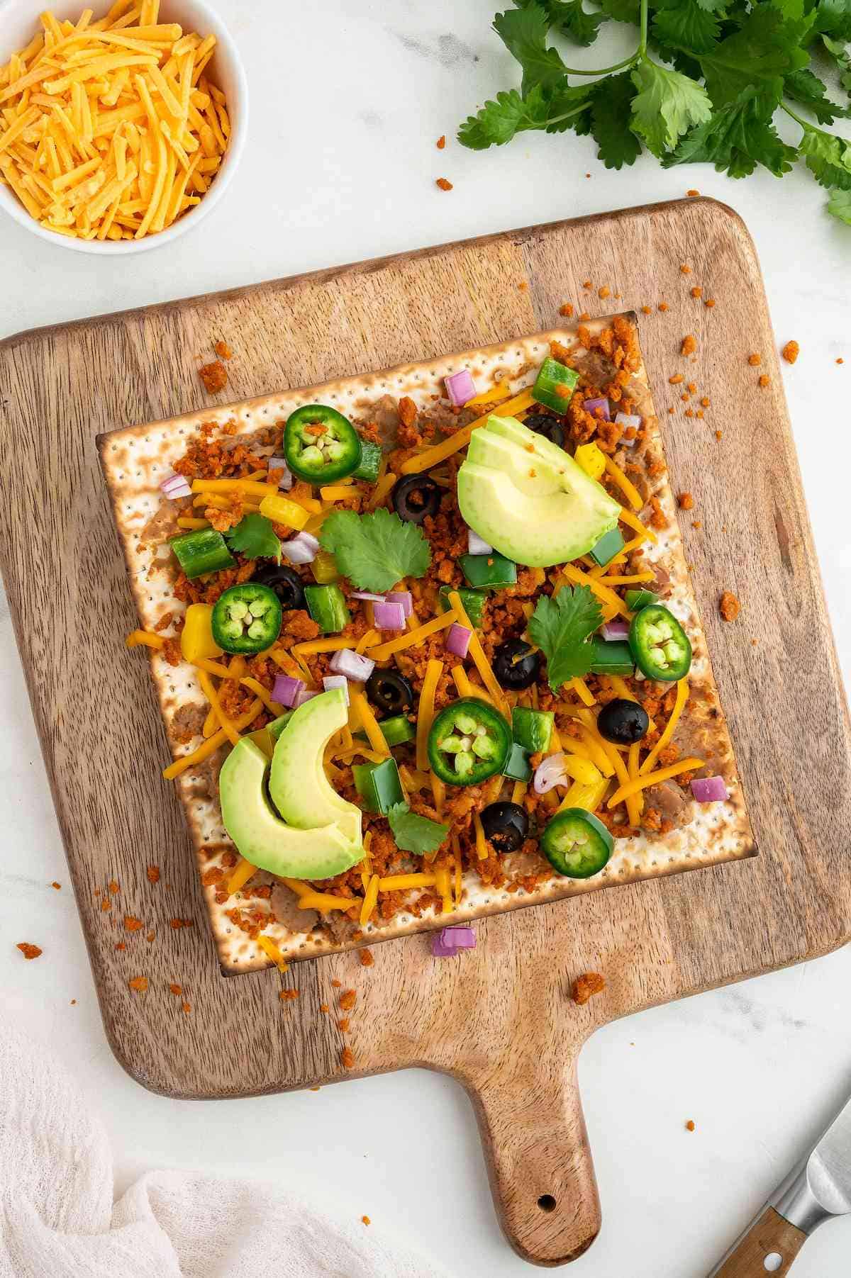 Matzah pizza topped with vegan refried beans, vegan cheese, and slices of jalapeno and avocado.