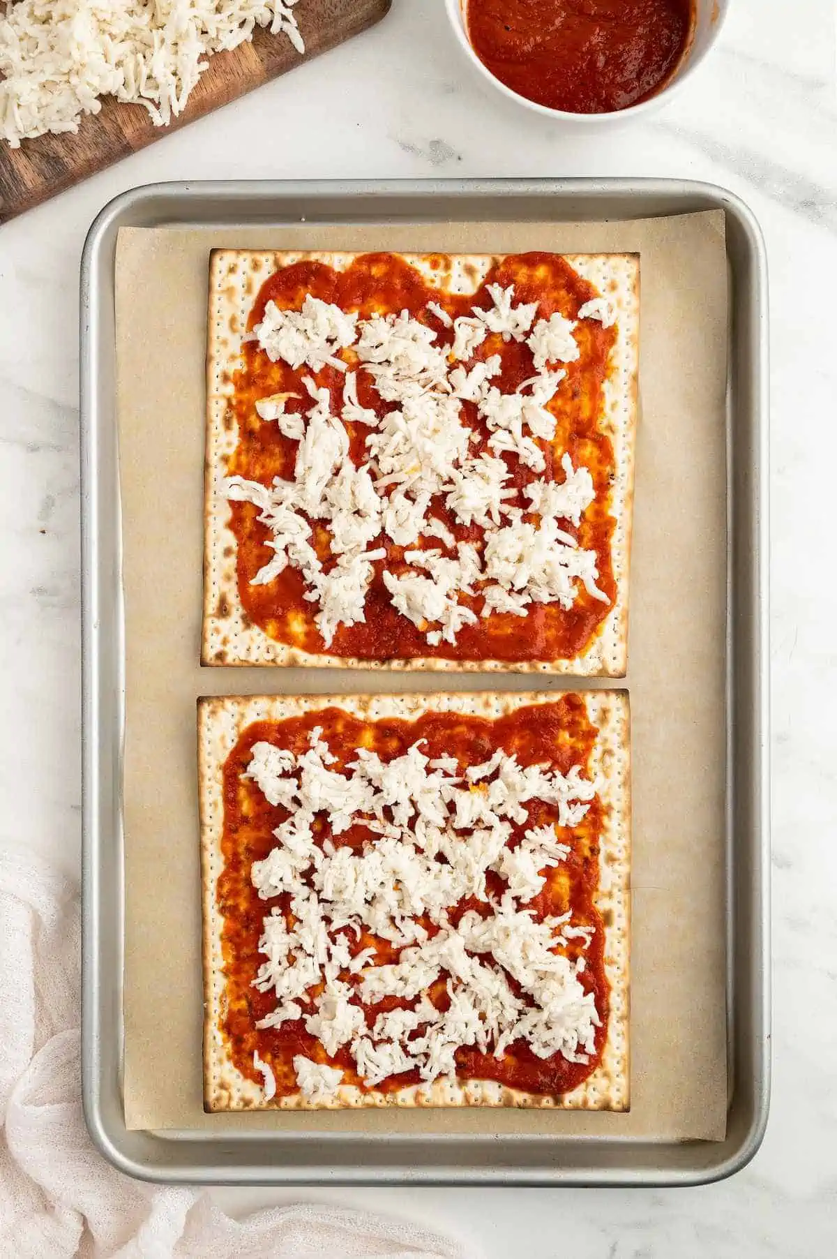 Two matzah crackers on a baking tray topped with pizza sauce and vegan shredded cheese.