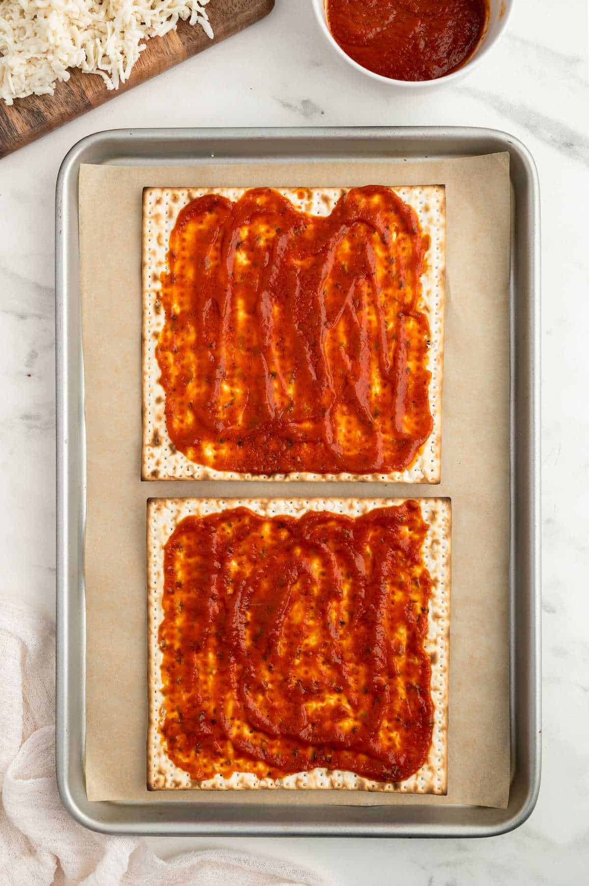 Two matzah crackers on a baking tray topped with pizza sauce.