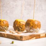 Homemade vegan caramel apples on sticks on a cutting board with pistachios.