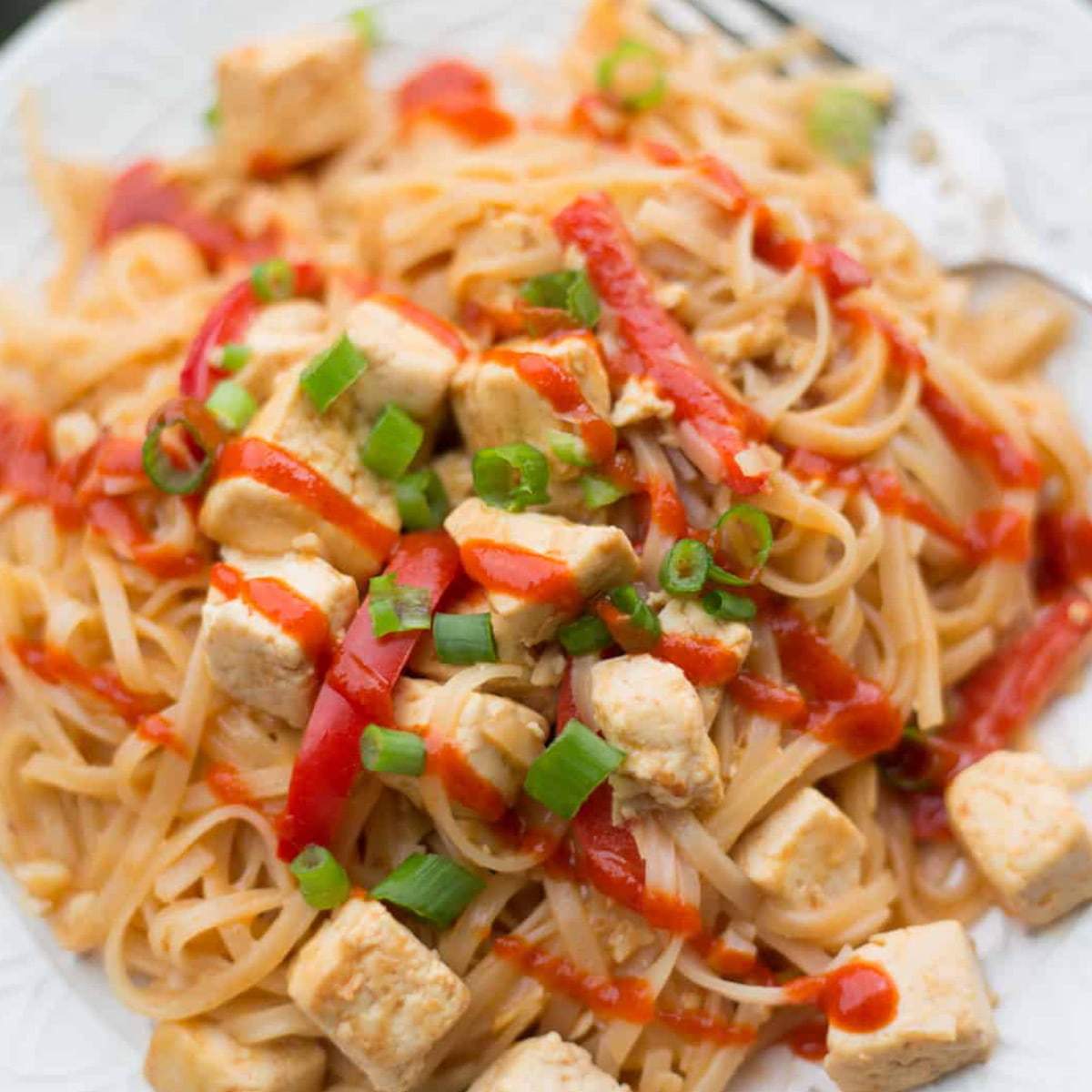 A plate of vegan pad thai topped with tofu, green onion, and a drizzle of Sriracha.