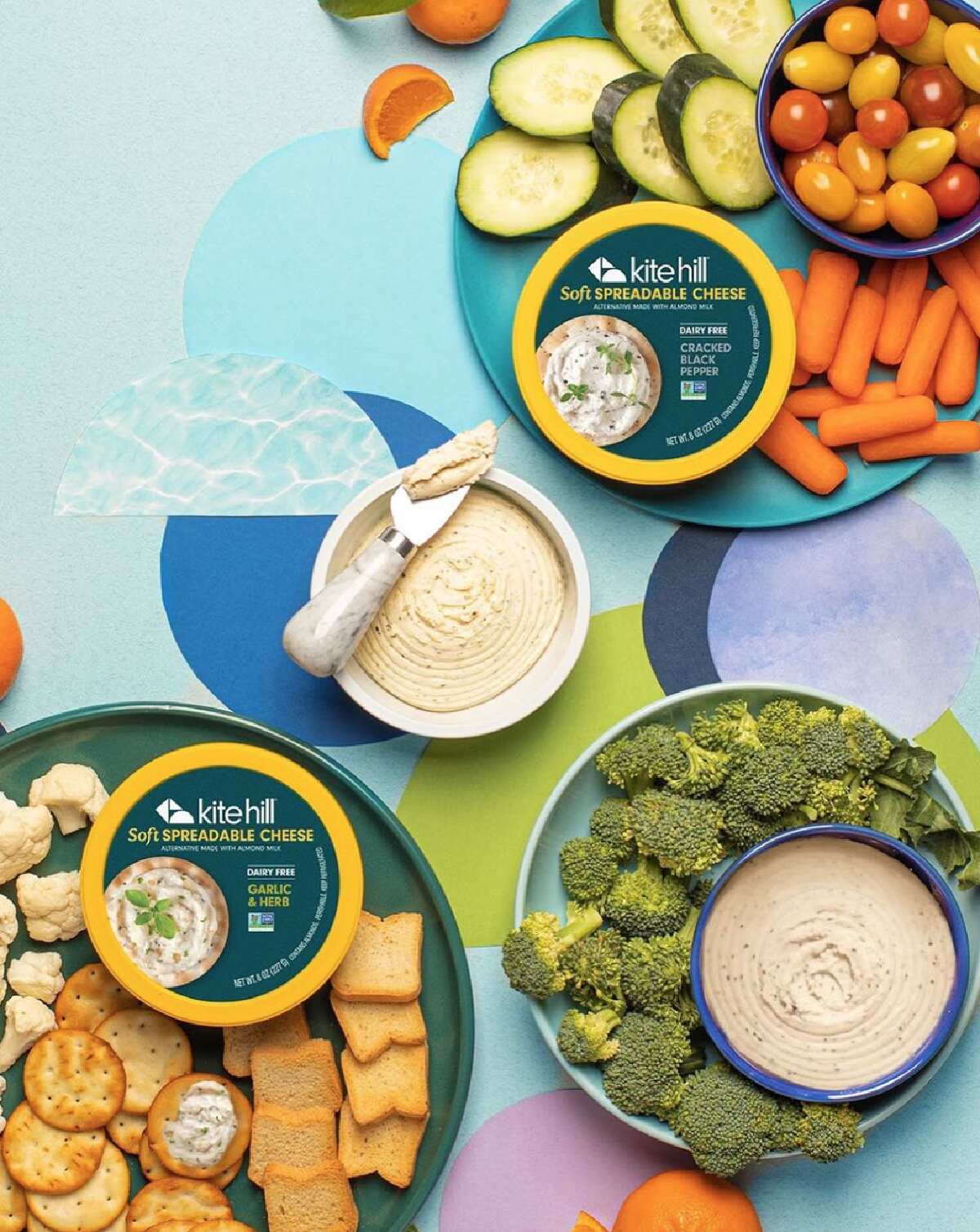 Two tubs of Kite Hill dairy free spreadable chese on colorful plates surrounded by crackers and vegetables on a blue and purple tablecloth background. 