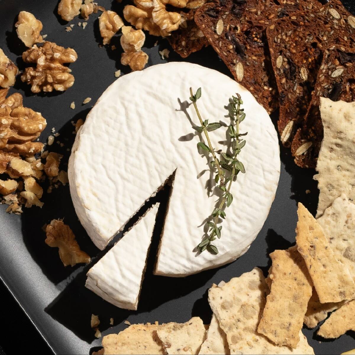 A round of Um Yum vegan cheese surrounded by crackers, cookies and nuts on a black platter. 