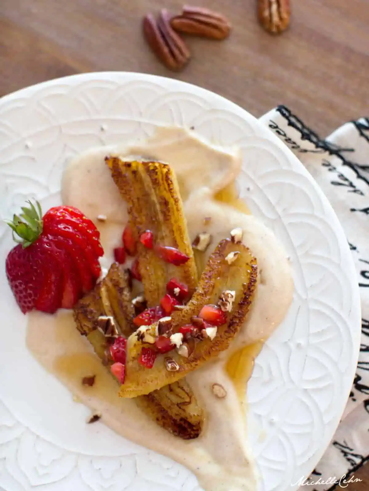 Pan fried cinnamon bananas on a plate with cream sauce and chopped strawberries and pecans.