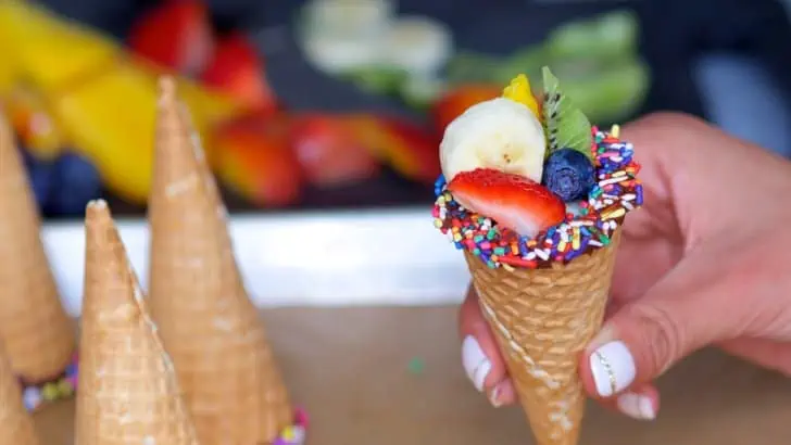 hand holding a chocolate-dipped cone with chocolate and sprinkles holding different fruits in it