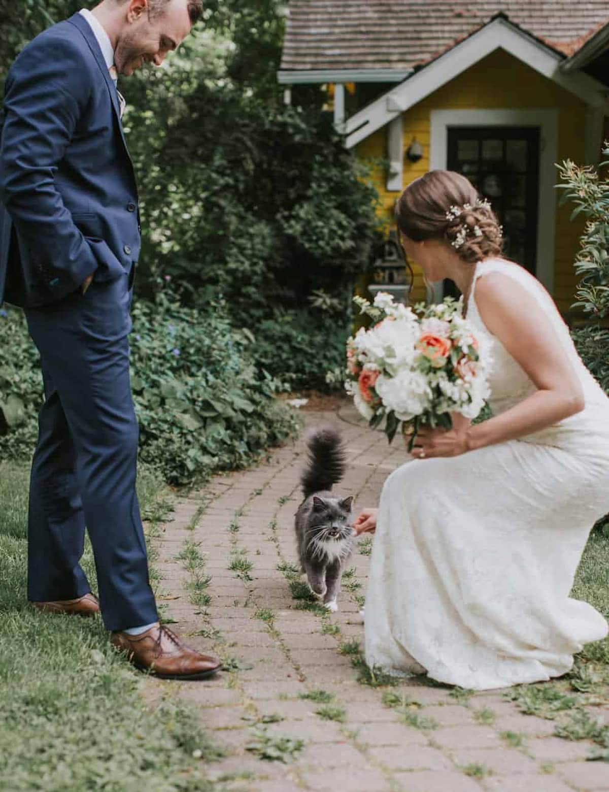Bride in a lace gown reaching out to a gray cat while the groom smiles nearby at their outdoor wedding.
