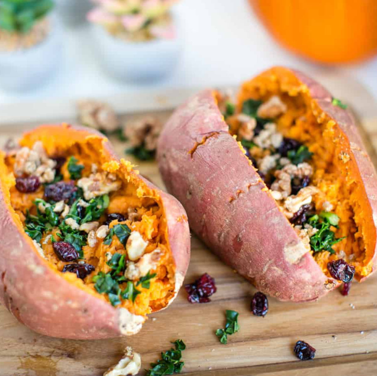 Two stuffed sweet potatoes on a cutting board, topped with candied walnuts, dried cranberries, and chopped kale.