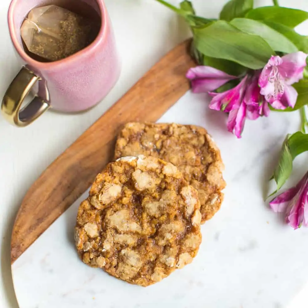 Two vegan oatmeal cookies on a marble serving platter next to fresh flowers and a mug of coffee.