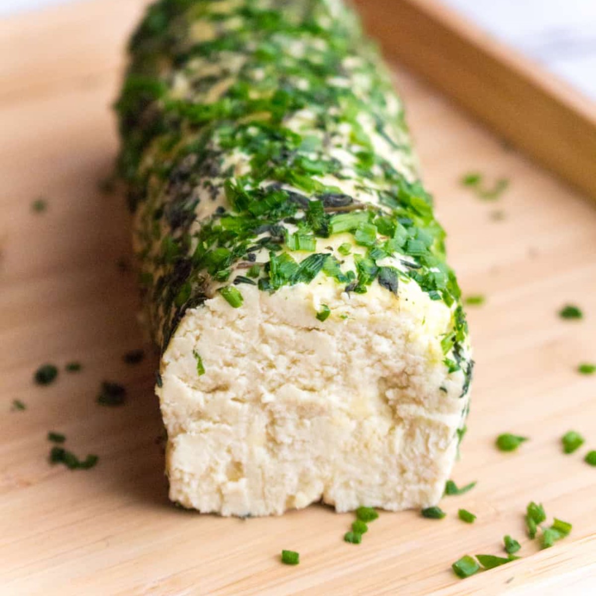 An almond-based vegan cheese log coated in sliced chives on a cutting board, with a slice taken out.