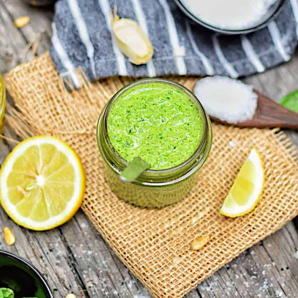 Vegan pesto sauce in a glass jar with spoon next to slices of lemon, garlic cloves, and a spoon of salt.