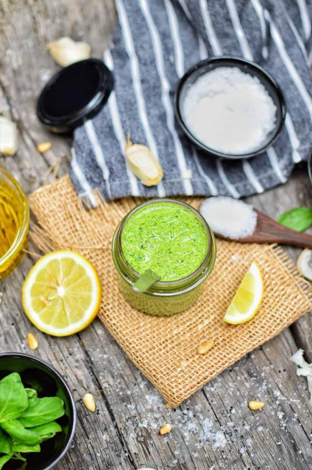 Vegan pesto sauce in a glass jar with various ingredients in bowls surrounding it.