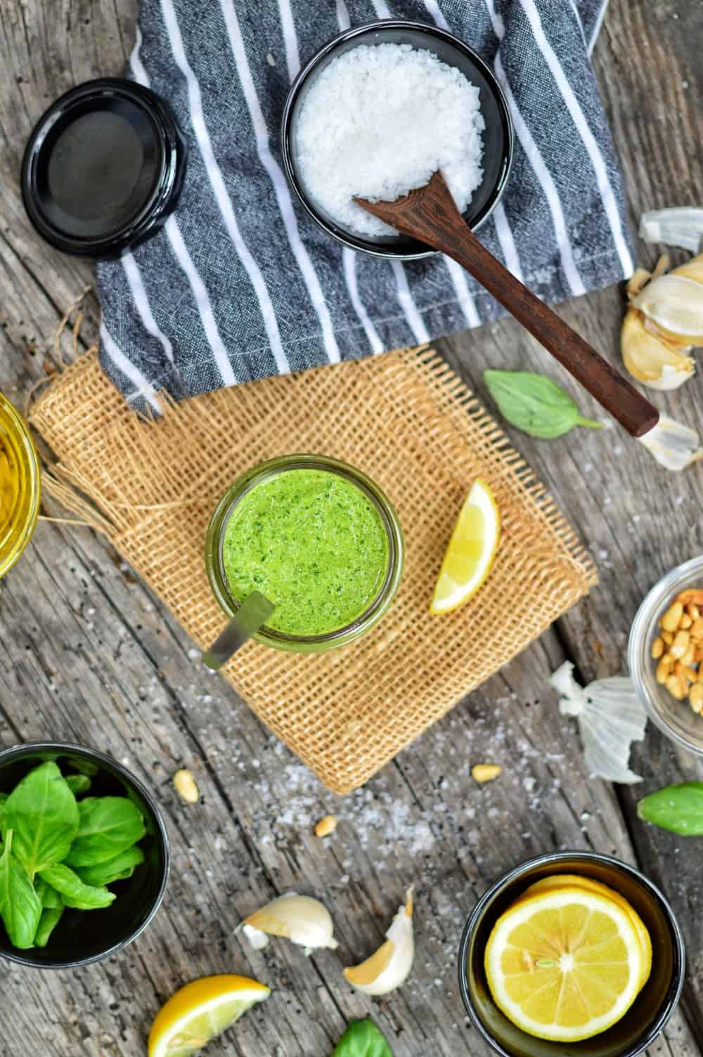 Vegan pesto sauce in a glass jar with various ingredients in bowls surrounding it.