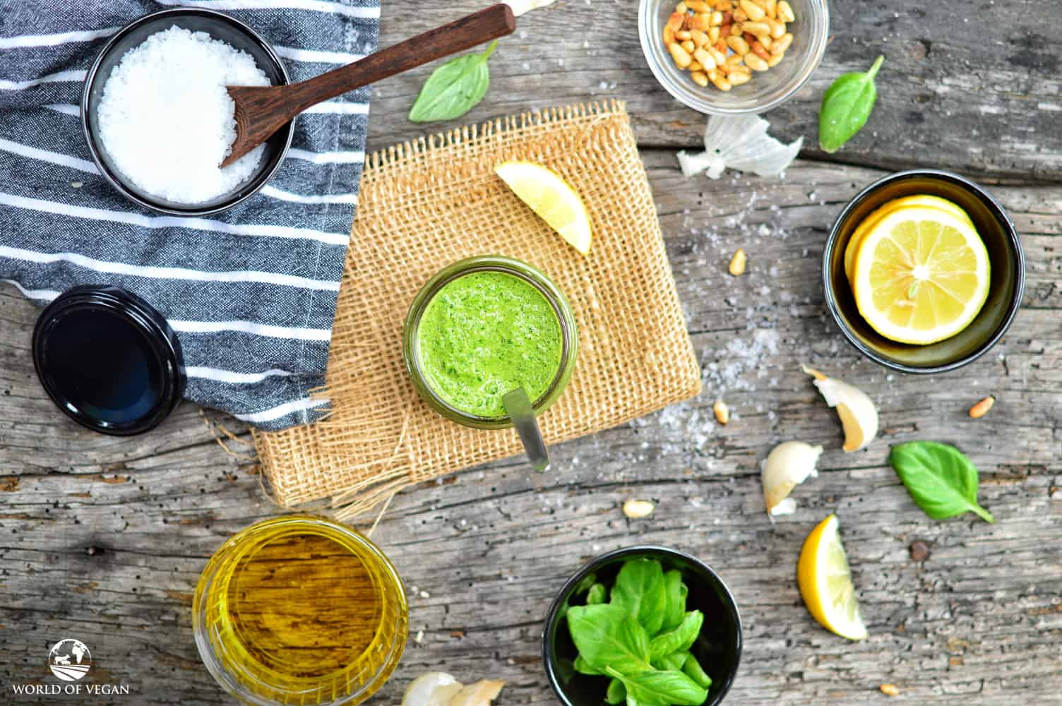 Vegan pesto sauce in a glass jar with various ingredients in bowls surrounding it.