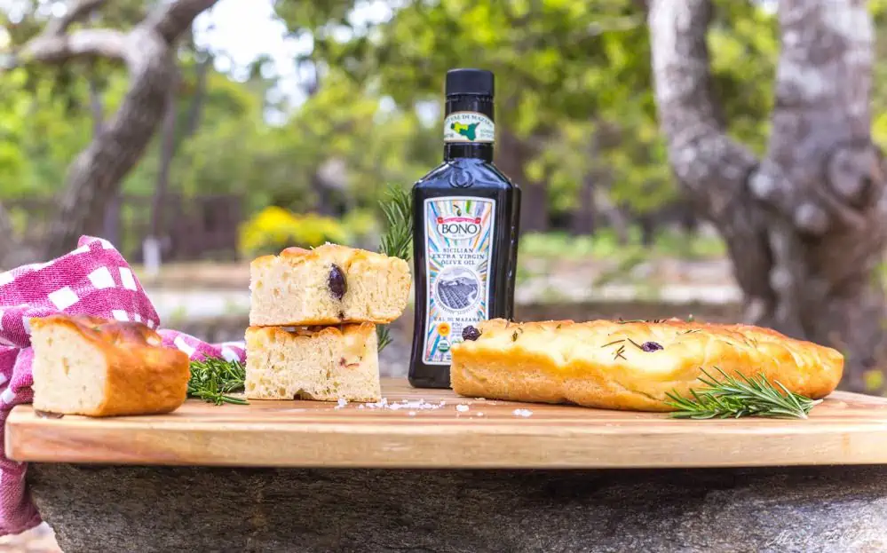 A wooden cutting board holding a bottle of Bono Olive Oil,  rosemary vegan focaccia bread, a red and white checked napkin and fresh sprigs of rosemary.