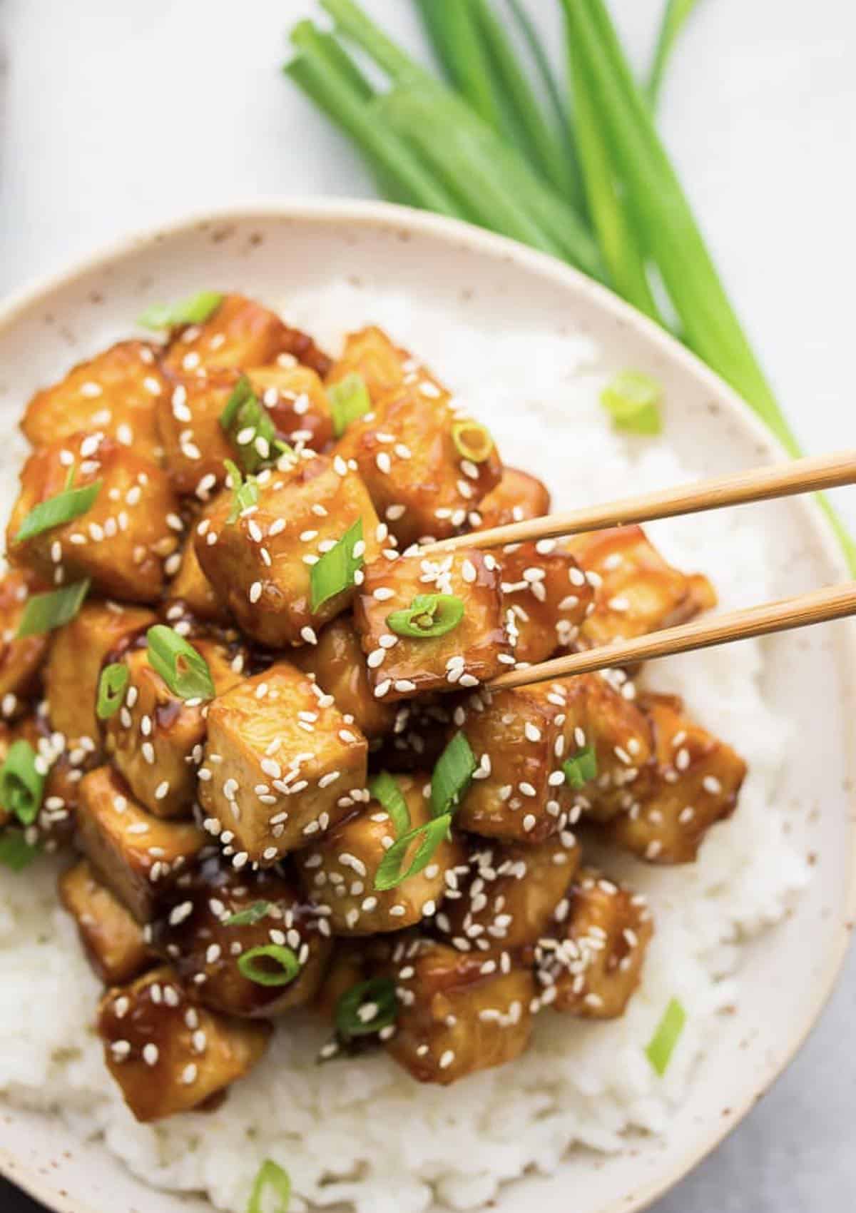 Plate of sticky sweet sesame tofu over white rice with chives and chopsticks. 