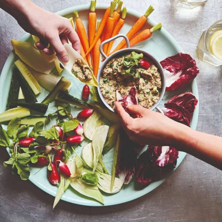 Hands reaching for baba ganoush dip on a vegetable platter.