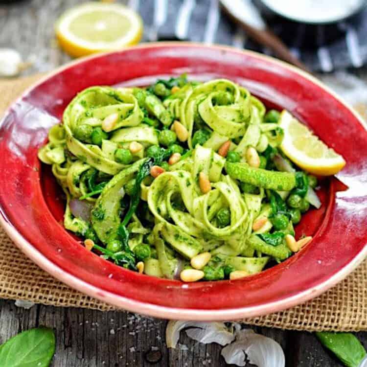 A bowl of vegan pesto pasta served on a cutting board surrounded by various pesto ingredients.