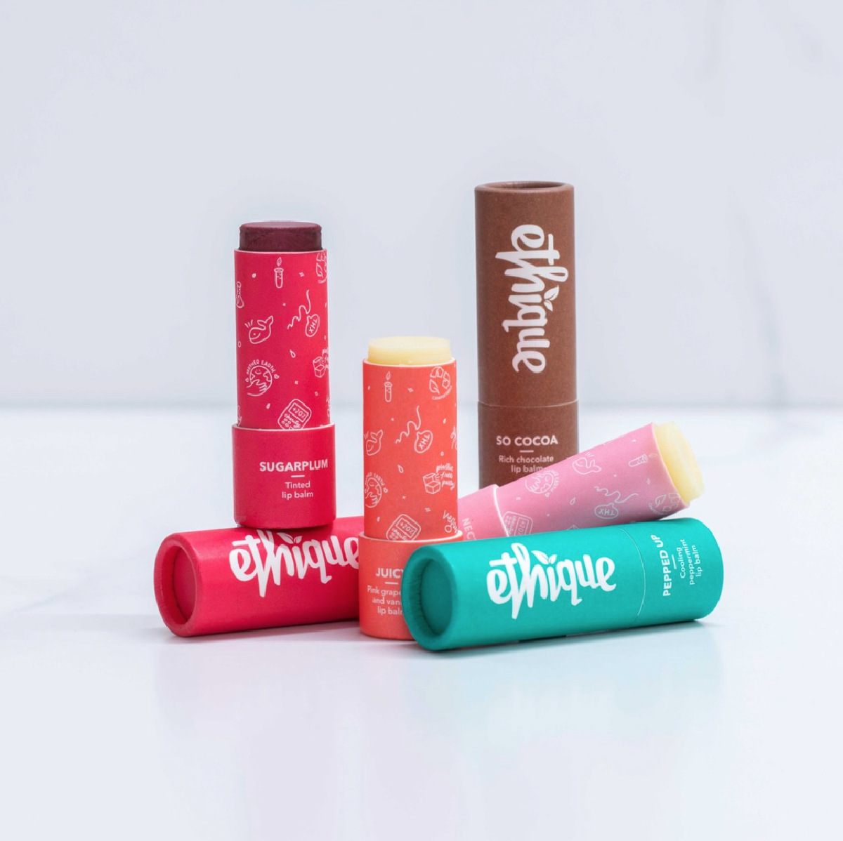 Five colorful eco-friendly tubes of Ethique vegan lip balms casually arranged on a white surface against a white background. 