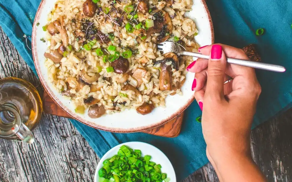 Plate of vegan mushroom risotto with a hand with pink fingernails holding a fork, ready to dig in.