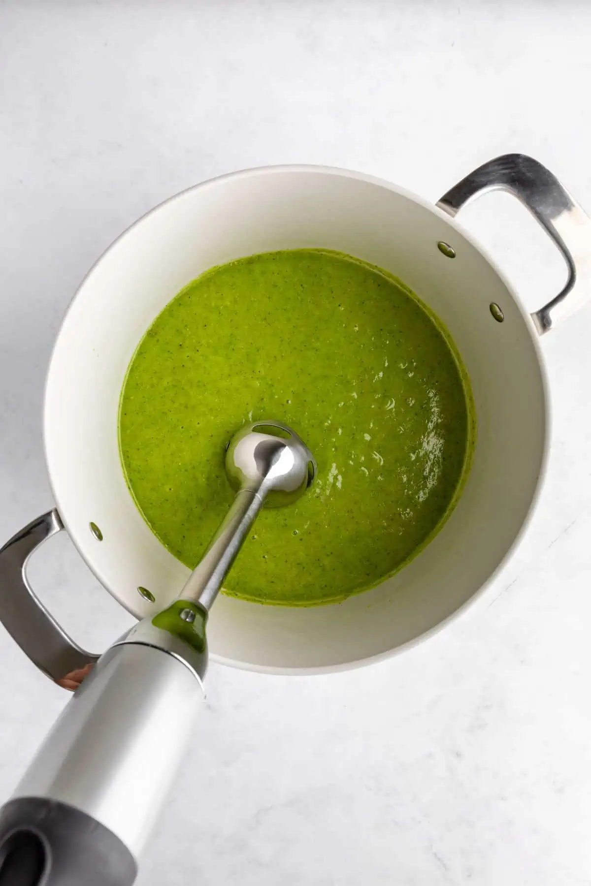 Blending the green pea soup with an immersion blender in a white pot.