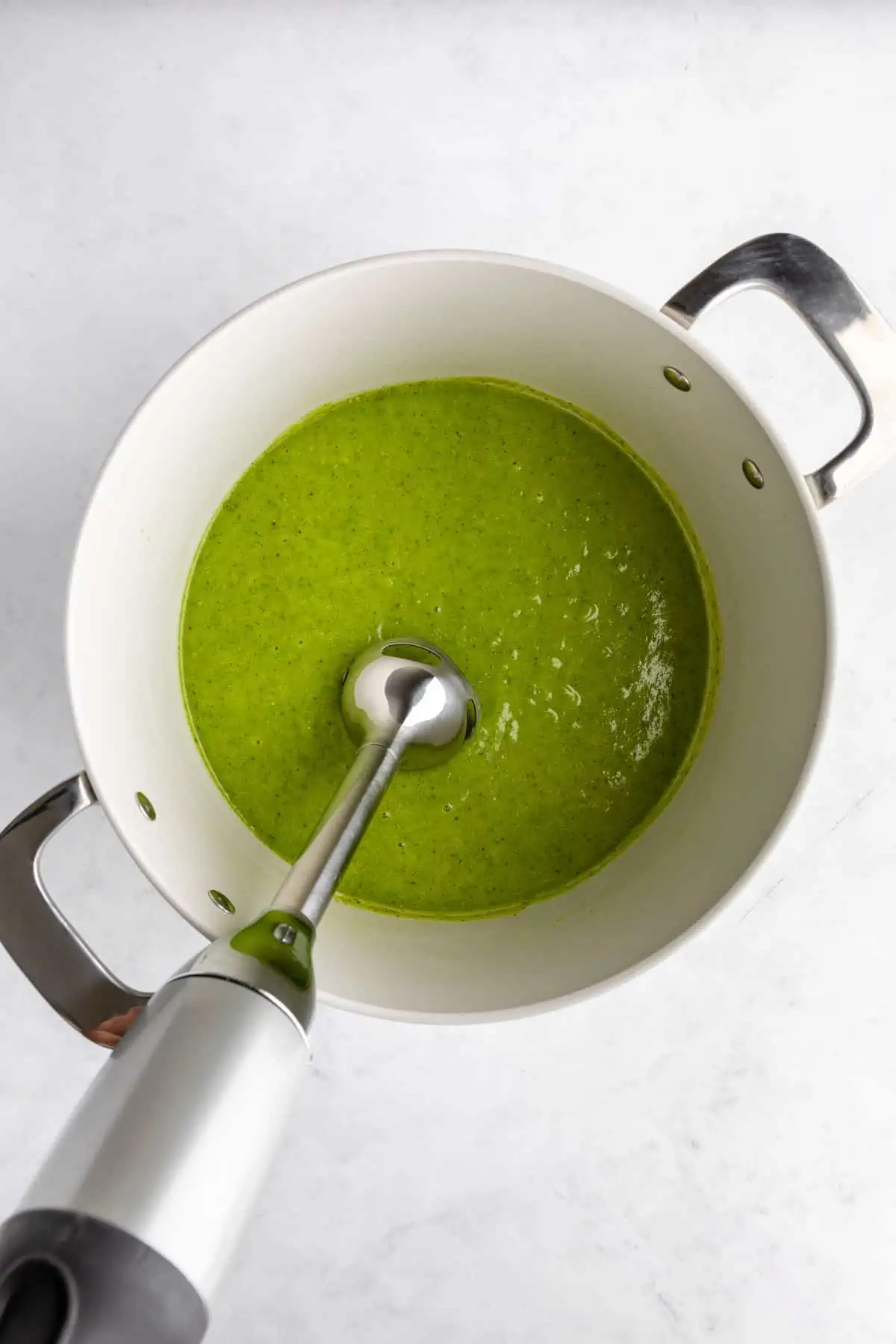 Blending the green pea soup with an immersion blender in a white pot. 