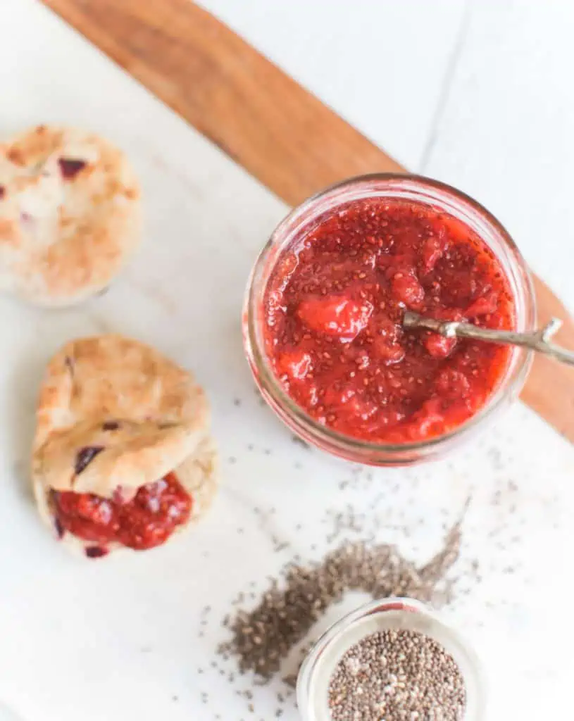 Vegan strawberry jam in a jar with a spoon, next to a jar of chia seeds and a biscuit smeared with jam.