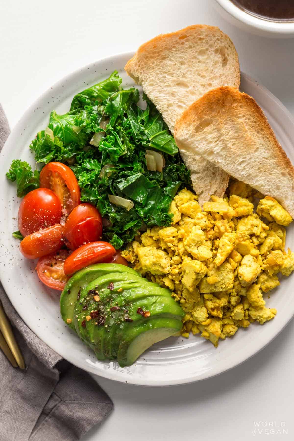 Vegan breakfast plate with tofu scramble, toast, avocado, tomatoes, and greens.