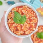A hand holding a bowl of vegan spaghettiOs made with dinosaur-shaped pasta.