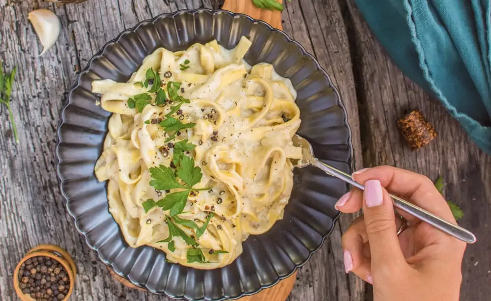 A plate of vegan fettuccine alfredo with a hand holding a fork.