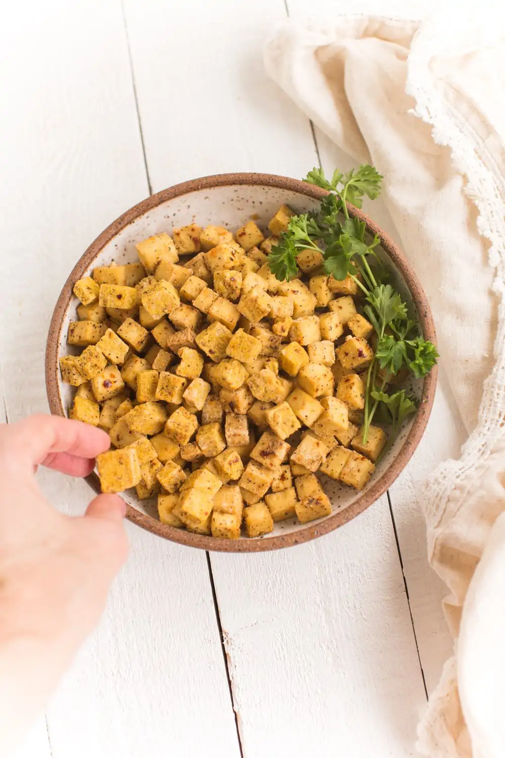 bowl of air fried tofu with parsley garnish and a hand on the left taking a piece