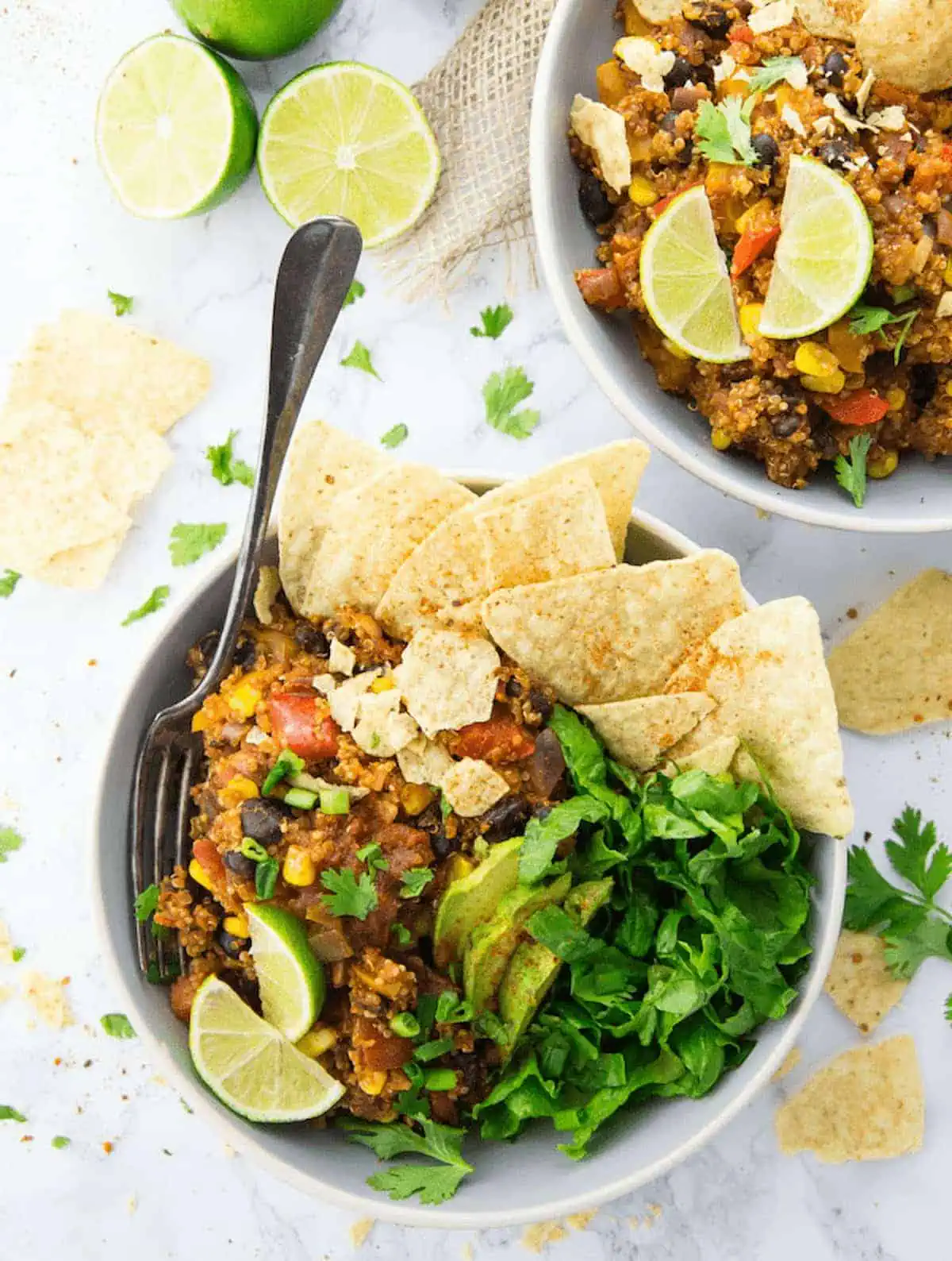 Two vegan burrito bowls with quinoa topped with lettuce, avocado, lime slices, and tortilla chips.