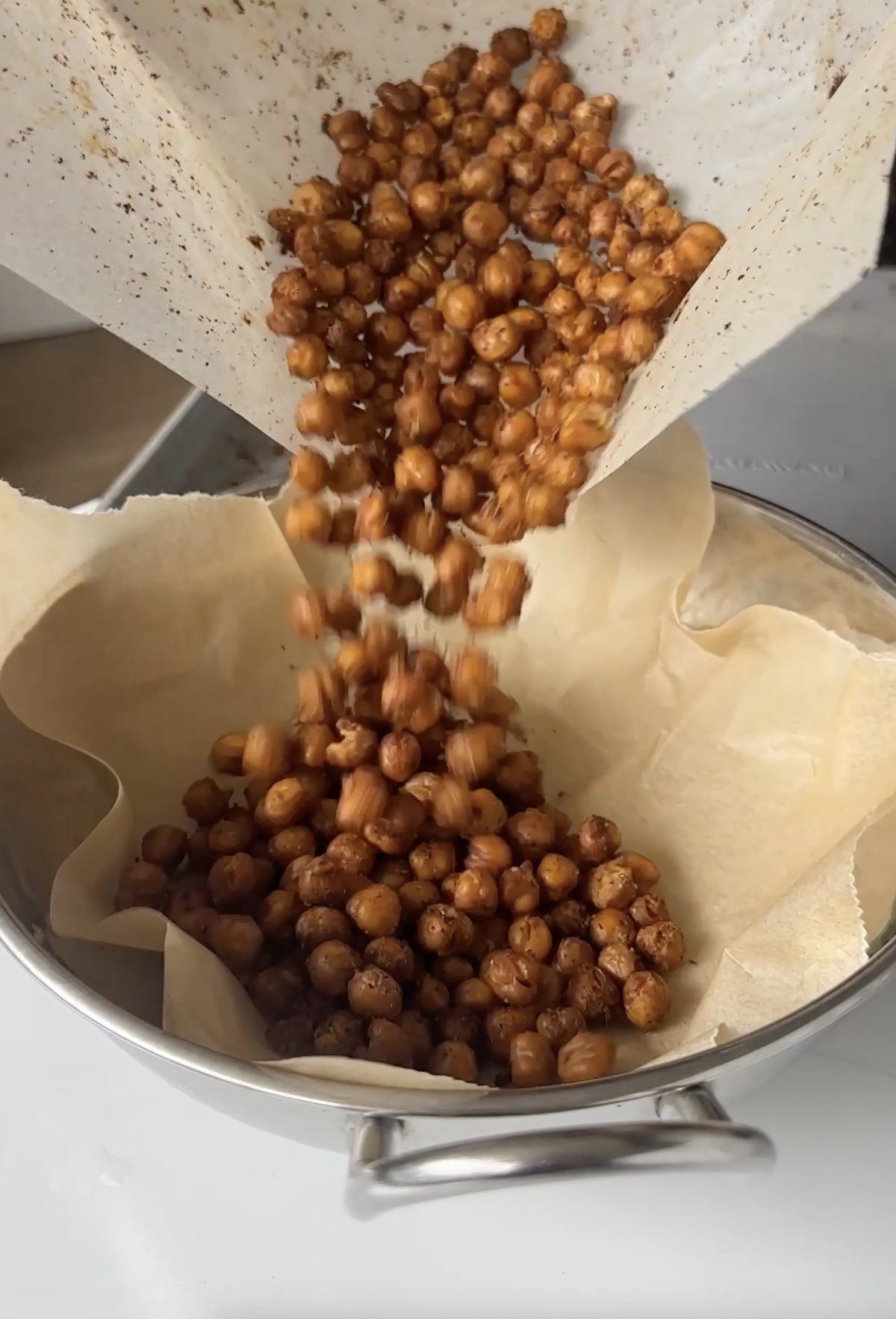Pouring crispy savory roasted chickpeas into a bowl lined with parchment paper.