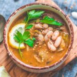 Vegan green chili soup with white beans, lemon slice, and parsley in a rustic bowl on a wooden board.