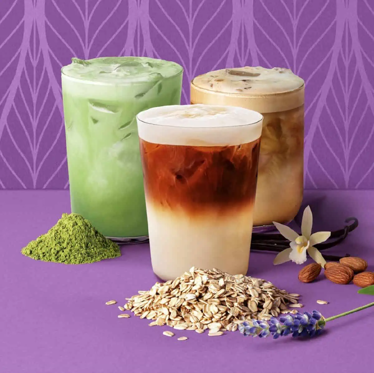 Three glasses of plant-based Peet's Coffee lattes for spring on a purple background with match powder, ground oats, vanilla flower, lavender and almonds on the counter next to appropriate drink. 