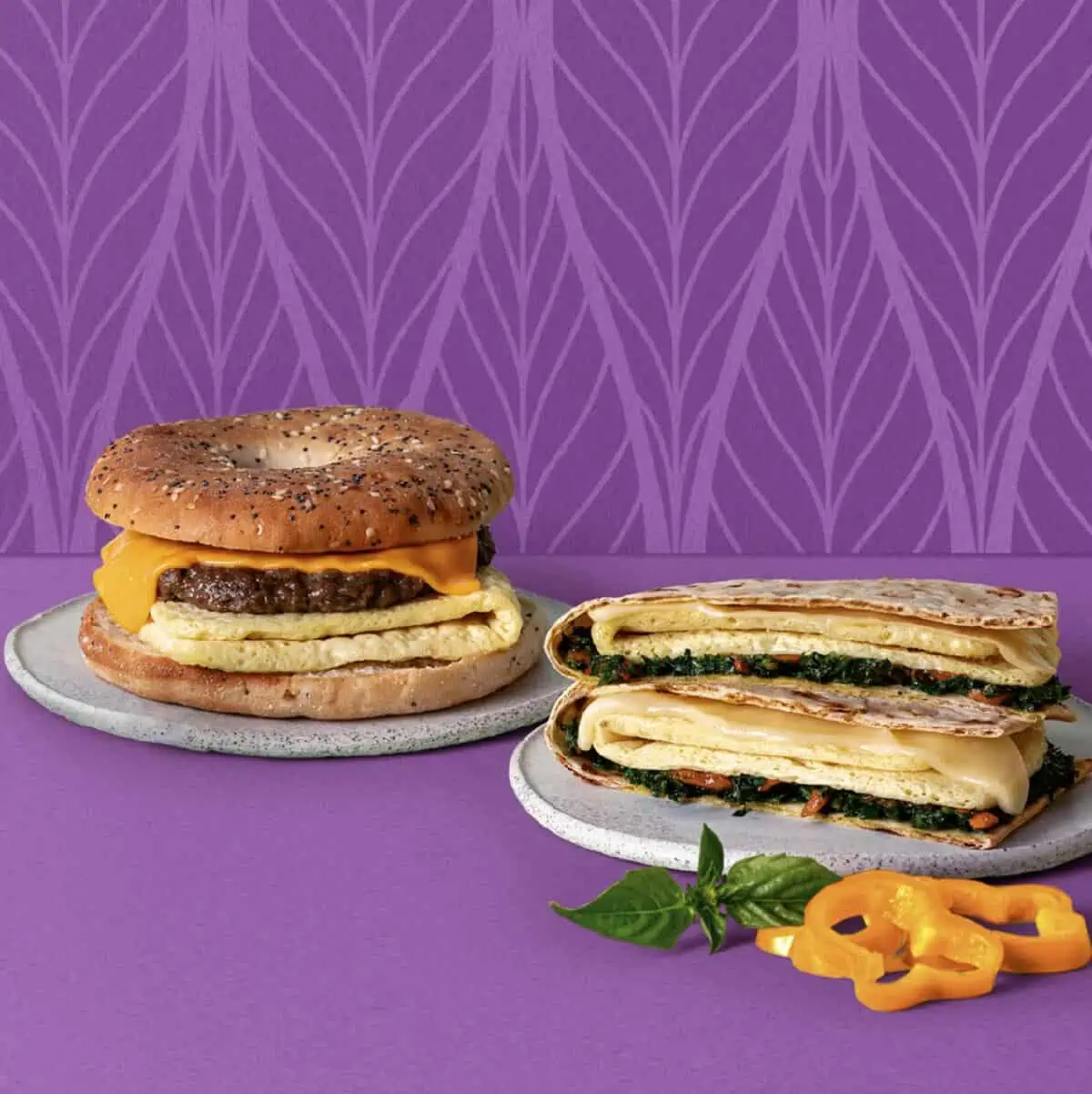 One vegan Just Egg and Violife Cheese Everything Bagel Sandwich and Mediterranean wrap laying on white plates on a purple patterned background with slices of orange bell pepper and spinach on the side.