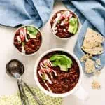 picture of chili in bowls on a light-colored table with blue cloths and measuring spoons around them. The chili is drizzled with cashew sour cream and slices of avocado on top.