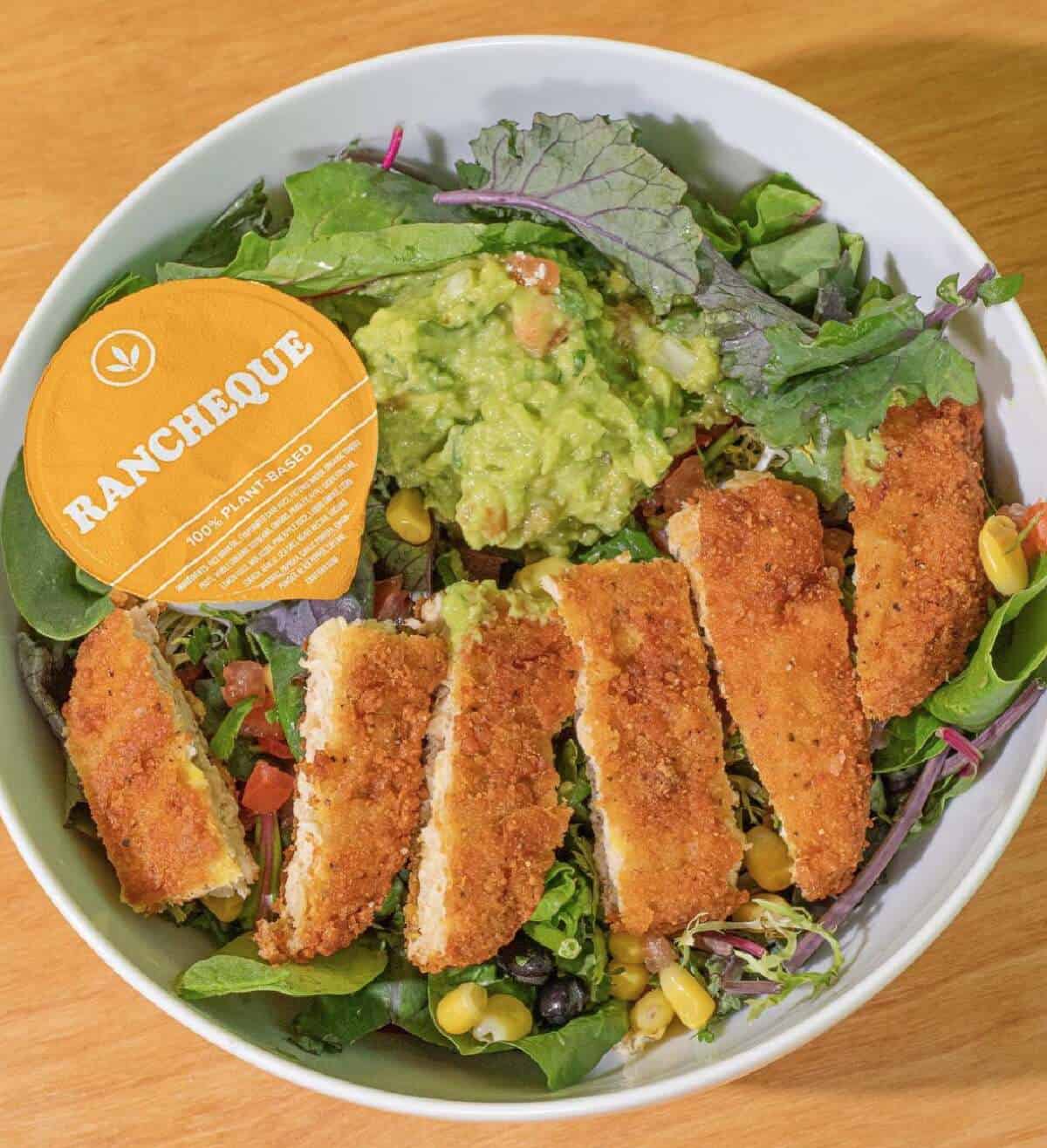 A white bowl filled with vegan chicken, salad, and a ranch/barbecue dressing on a wooden countertop.