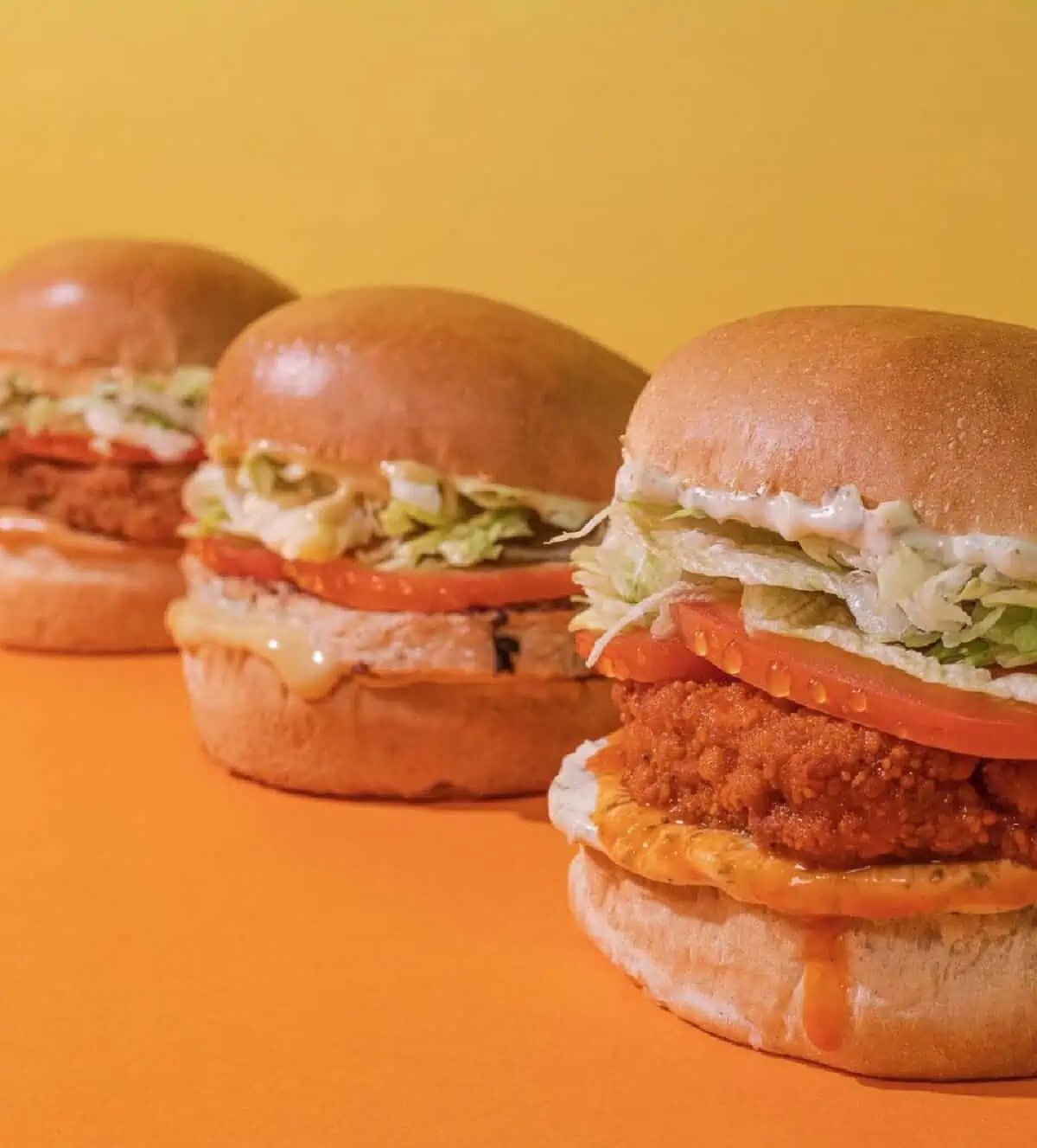 Three Plant Power Fast Food 'Chicken' Sandwiches in a diagonal row on an orange countertop with a background of yellow walls.