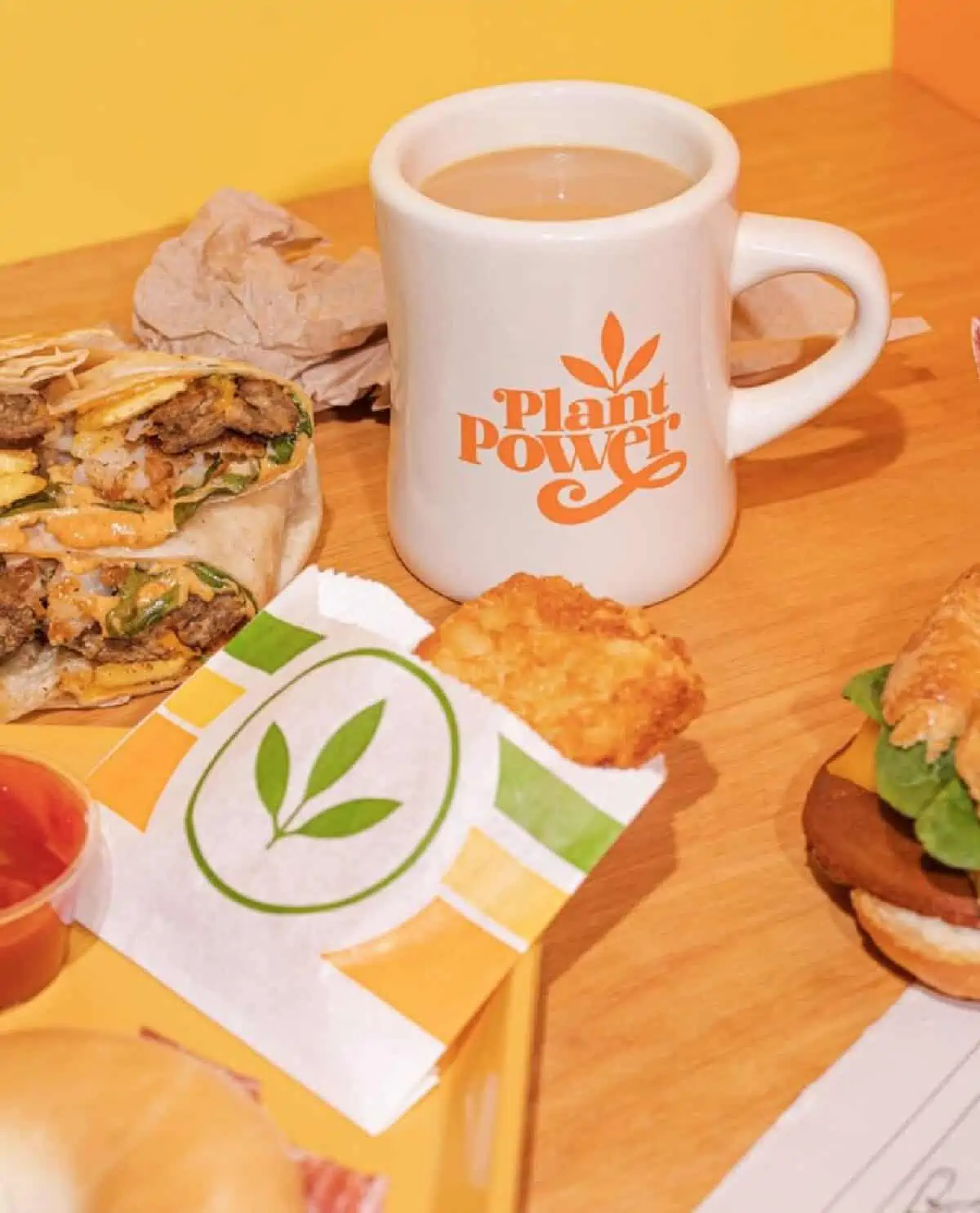 Plant-Based breakfast wraps, croissants, and a hash brown patty in a paper sleeve laying on a wooden countertop along with a vintage style white coffee mug full of coffee.