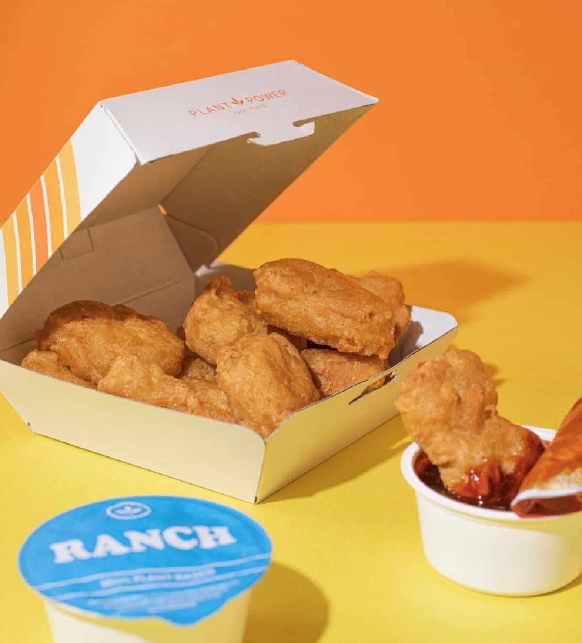 A Plant Power Fast Food cardboard box filled with 'Chicken' nuggets and two containers of dipping sauce, Ranch and Barbecue on a yellow countertop with background of orange walls.