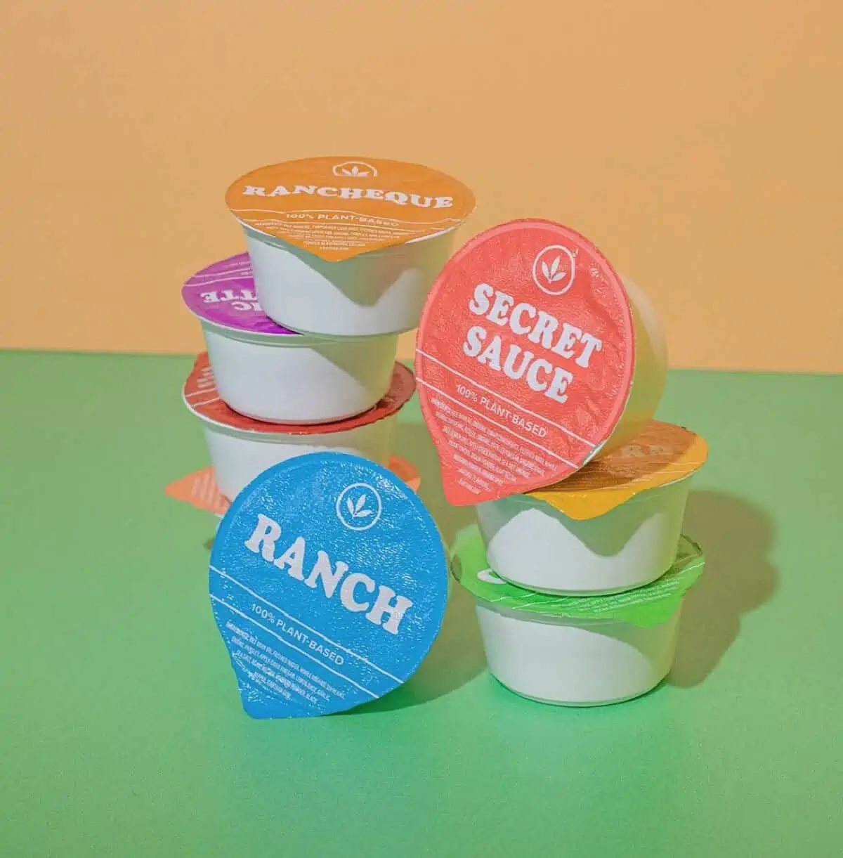 A stack of eight colorful circular plant-based dips and dressings on a green tabletop with an orange background.
